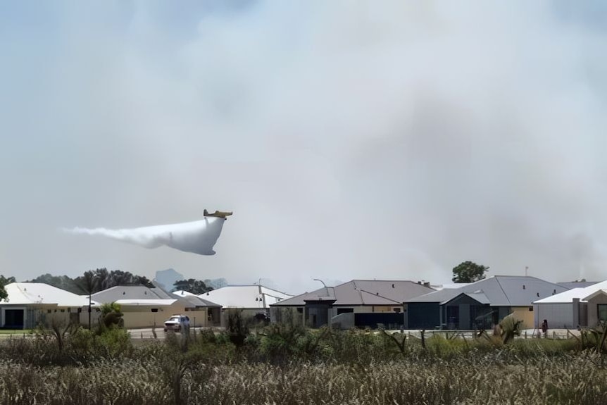 Fire fighting aircraft drops water over suburban houses with smoke in the background