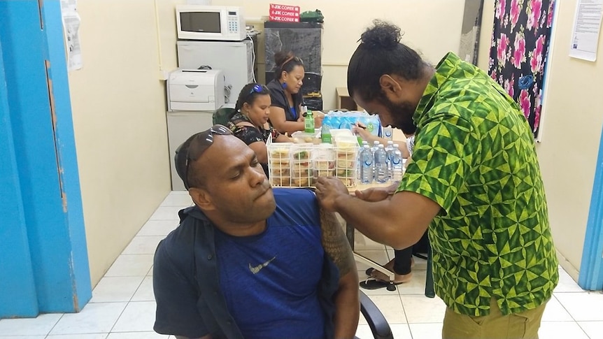 A man sitting down gets an injection from another man wearing a bright green shirt. 