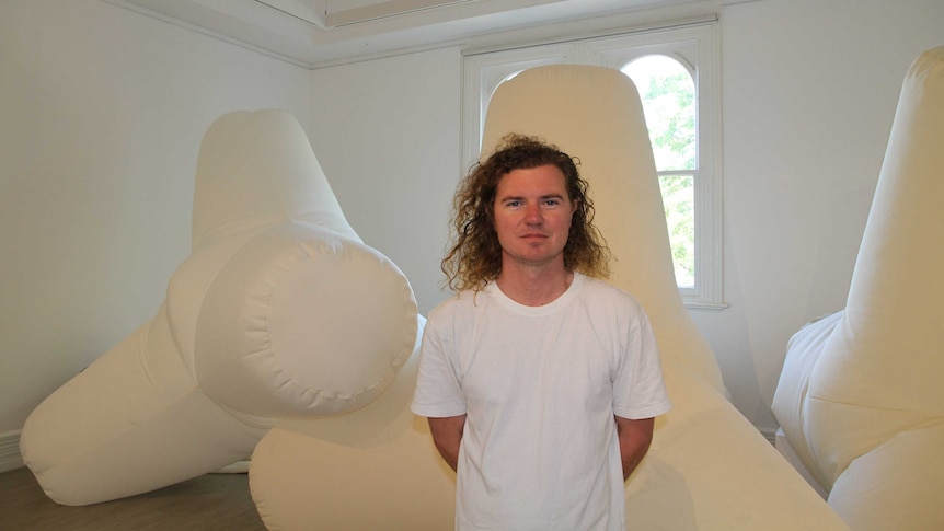 Man in white short sleeve tee stands with his hands behind his back. Behind him there are large fabric inflated shapes.