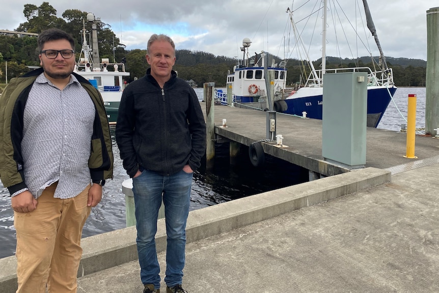 Two men stand on a concrete boat harbour in front of boats.