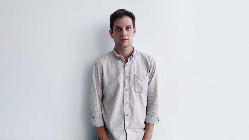 Evan Gershkovich wears a white shirt with sleeves rolled up as he stands in front of a white wall and looks neutrally ahead.