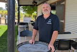 Bruce Heward, a middle-aged man with a beard and grey hair, stands in front of a wine barrel outside a corrugated iron bar.