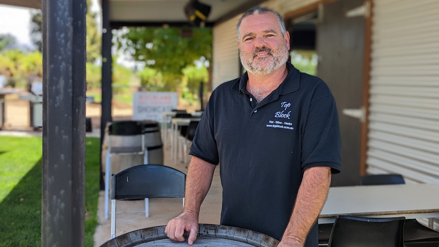 Bruce Heward, a middle-aged man with a beard and grey hair, stands in front of a wine barrel outside a corrugated iron bar.