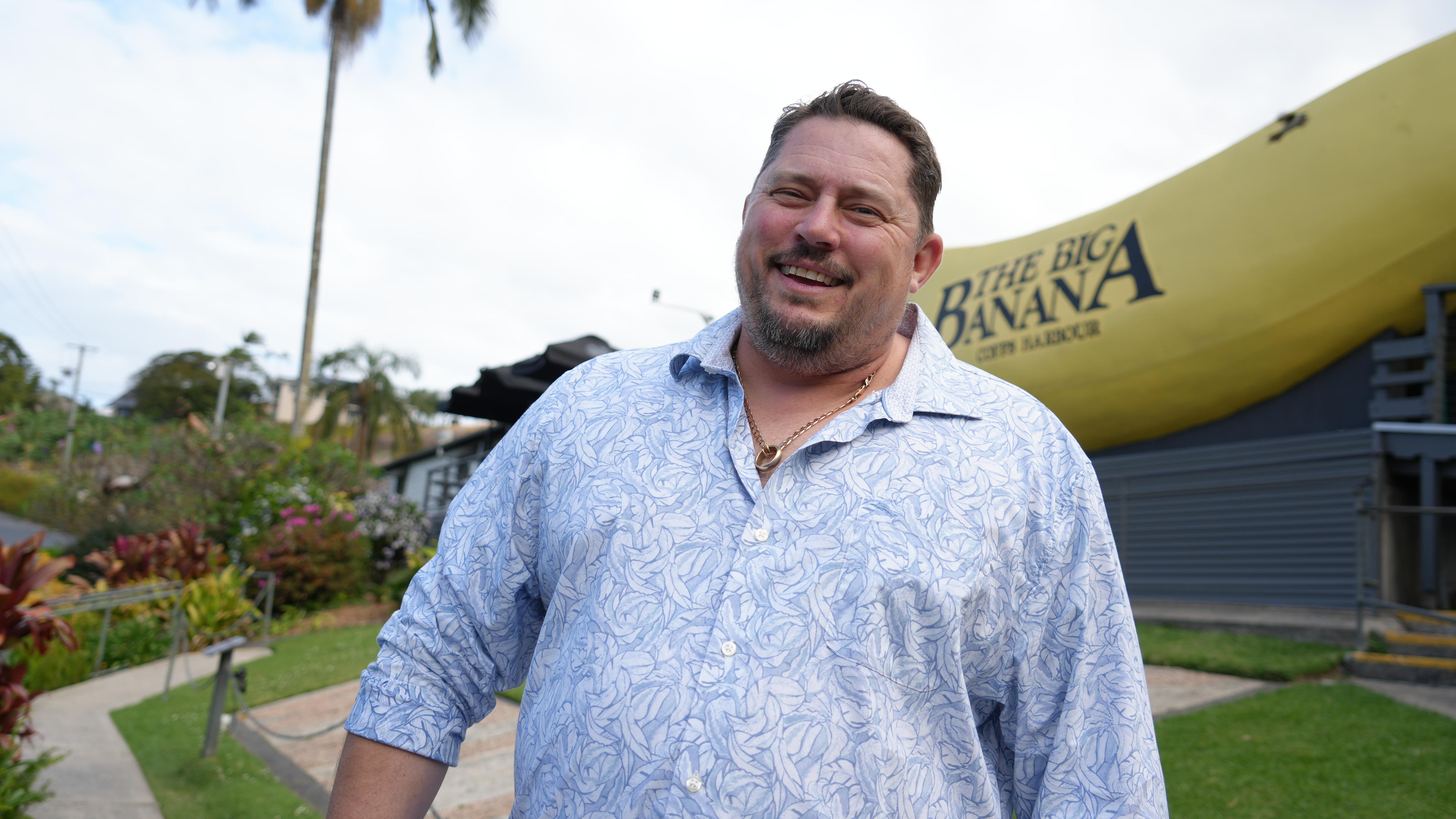 A smiling, bearded man in a collared long-sleeved shirt stands in front of the Big Banana.