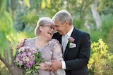 Couple in wedding outfits looking into each other's eyes with greenery in background.