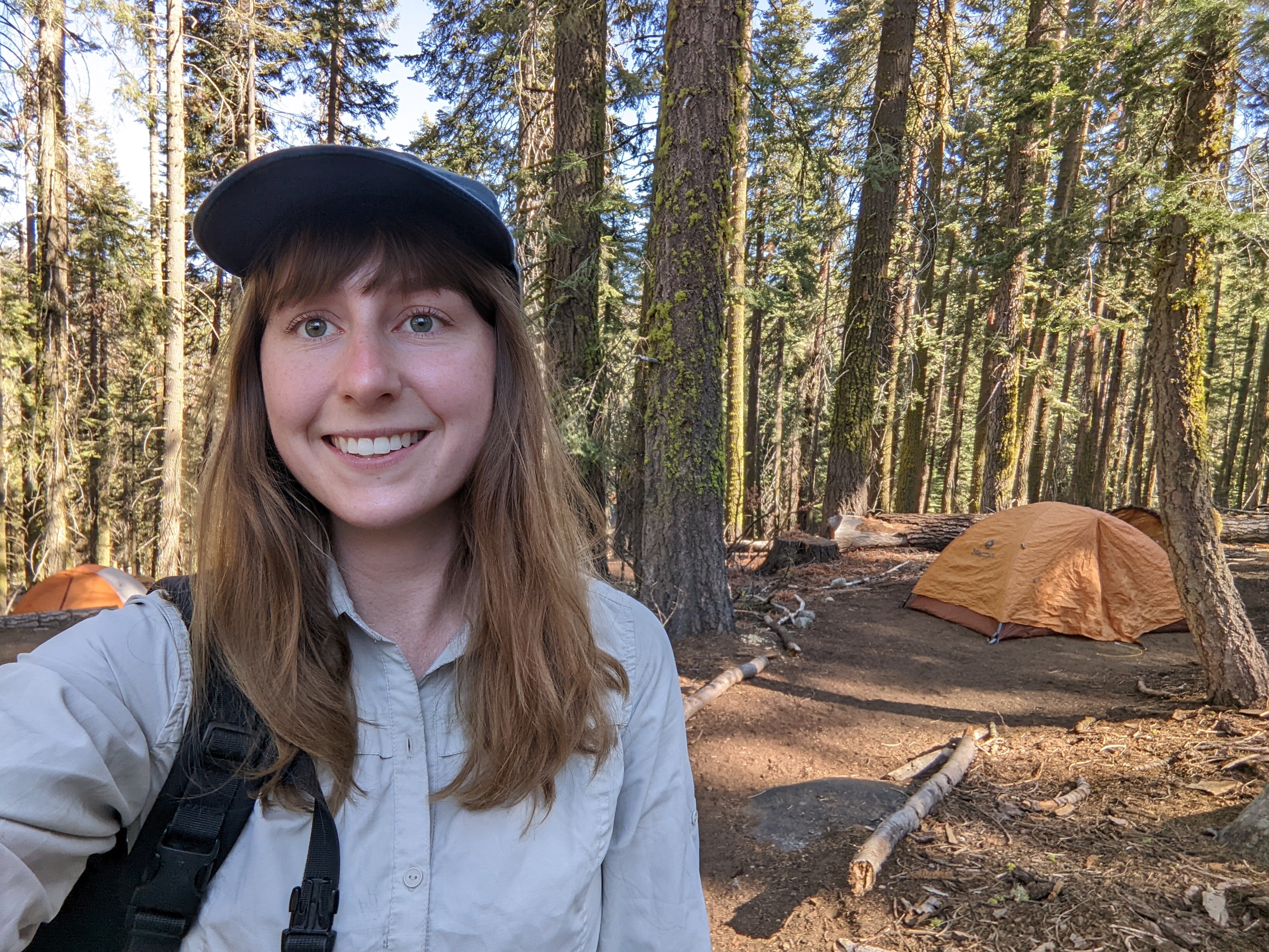 A young woman with light brown hair wears a blue cap and grey shirt, and smiles, with a forest behind her.