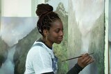 Melanie McCollin-Walker holds a small paintbrush in front of a large painting of a landscape.