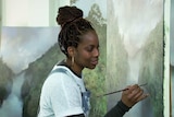 Melanie McCollin-Walker holds a small paintbrush in front of a large painting of a landscape.