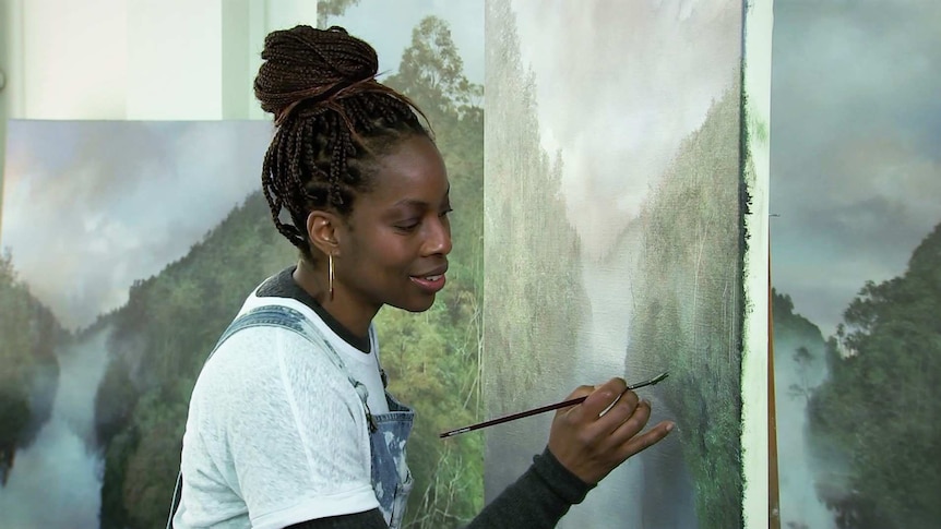 Melanie McCollin-Walker holds a small paintbrush in front of a large painting of a landscape.