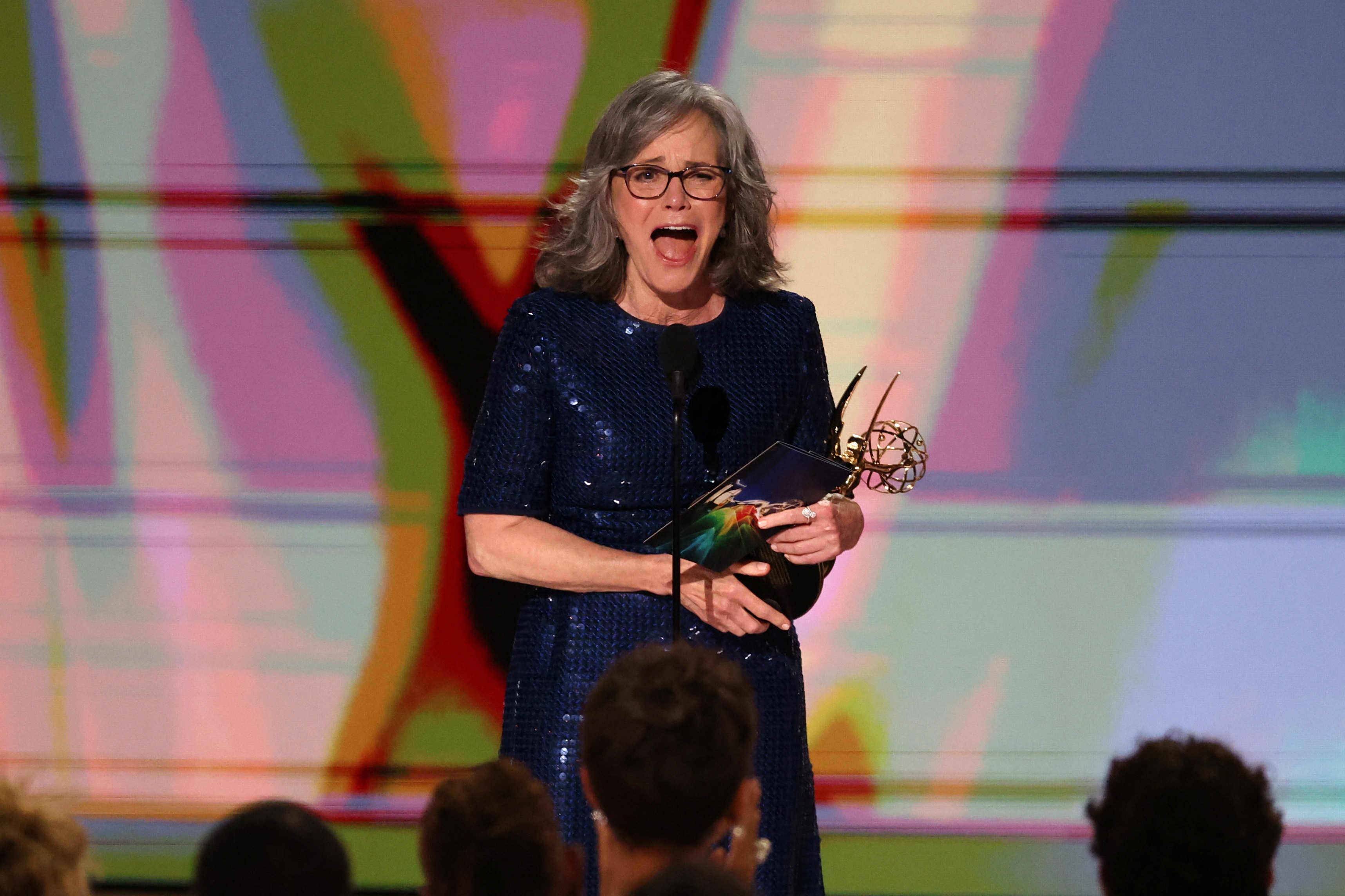 a woman in a blue sequen dress smiles at a mic stand holding an emmy statue