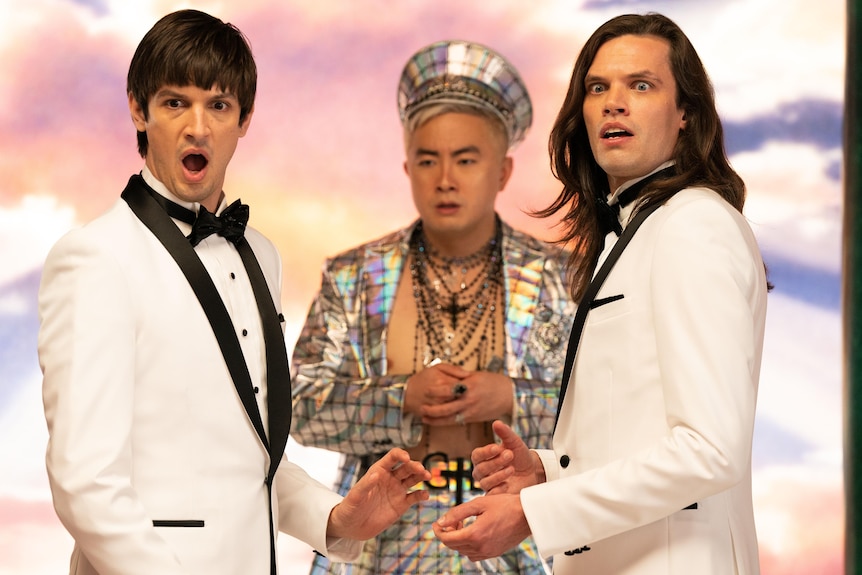 Josh Sharp and Aaron Jackson wear matching white tuxedos in front of God and heaven, with shocked faces at the camera.