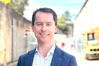 A man with short brown hair wearing a blue jacket over a white short. He is smiling.