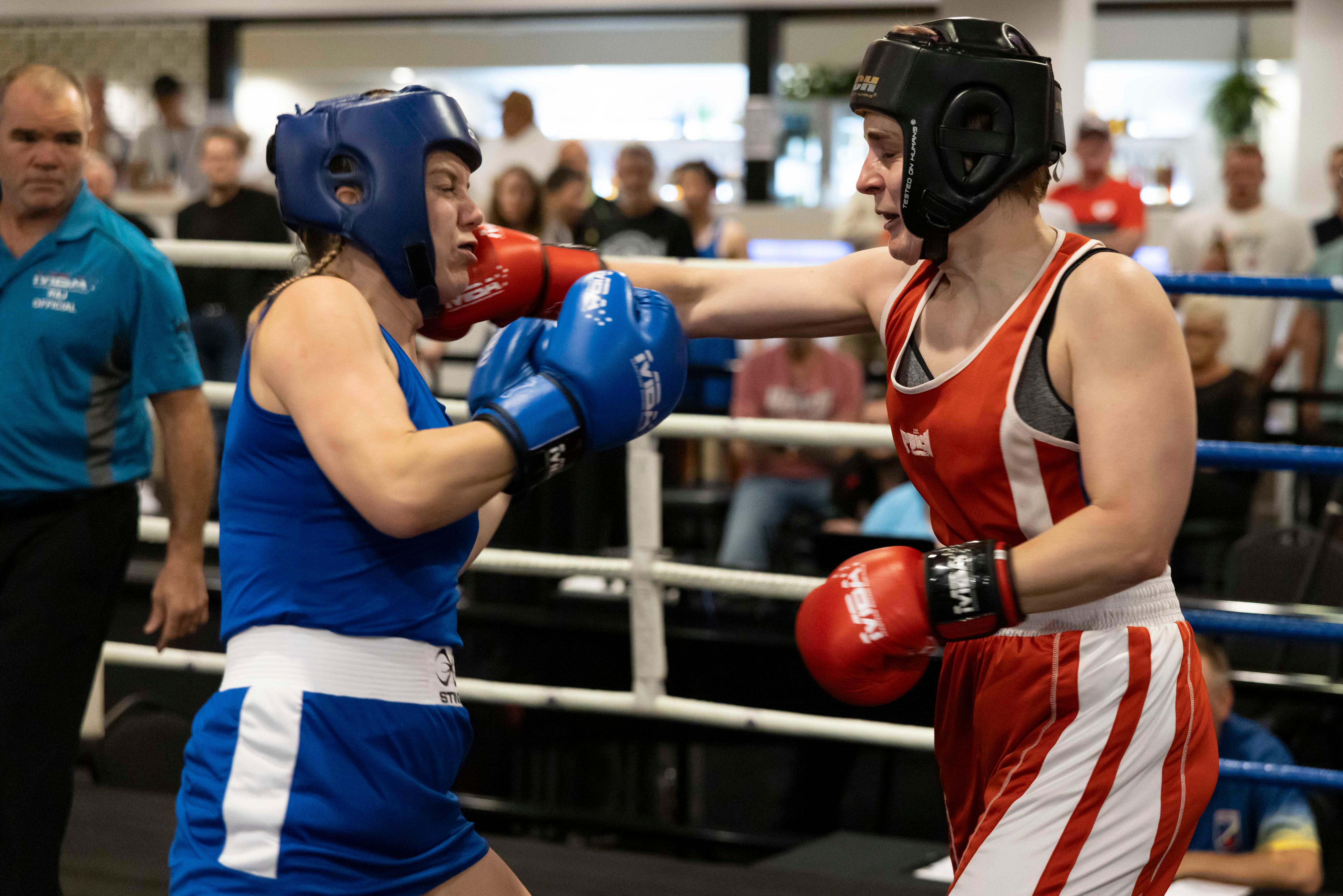 Duas mulheres em uniformes esportivos vermelhos e azuis no ringue de boxe com luvas de boxe vermelhas e azuis e uma multidão e um árbitro atrás delas
