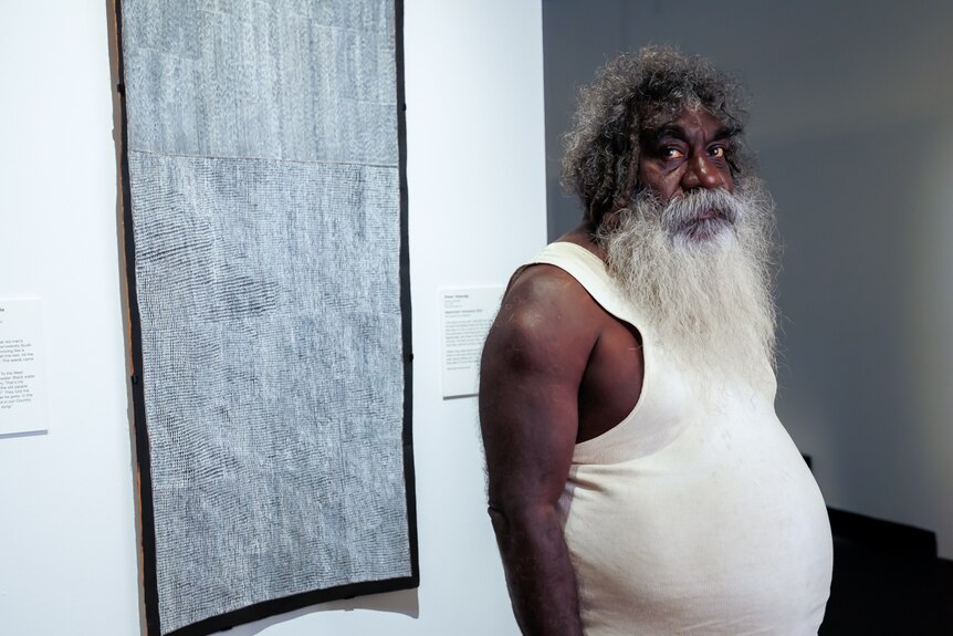 An Aboriginal man with a long beard stands in front of a black and white bark painting on a gallery wall
