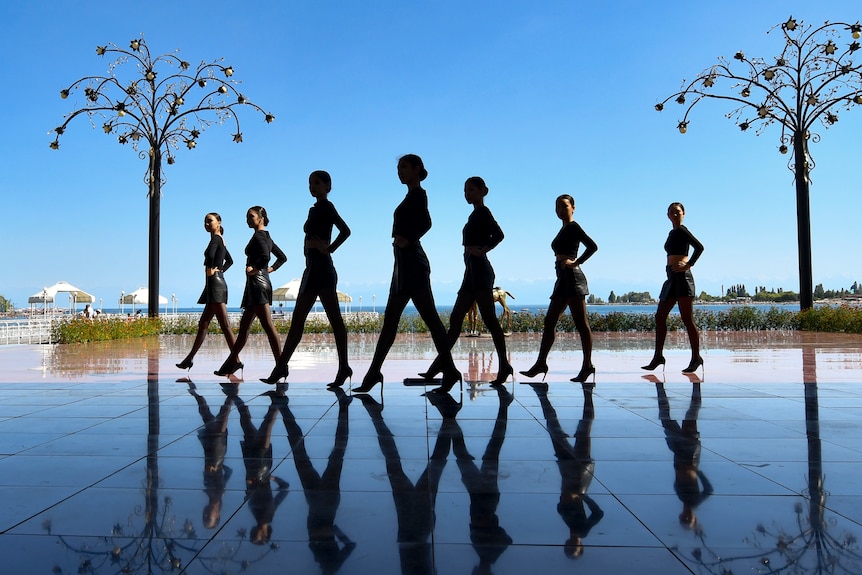 Silhouettes of seven female dancers against blue tiles and sky. 
