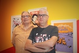 Two aged men stand in front of a bright wall filled with gig posters from the 80s