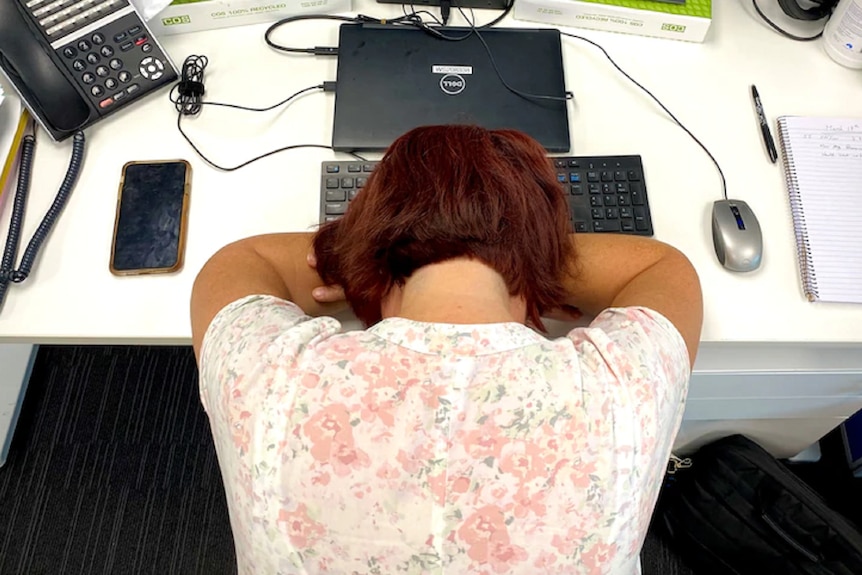 A woman rests her head on her folded arms on an office desk.