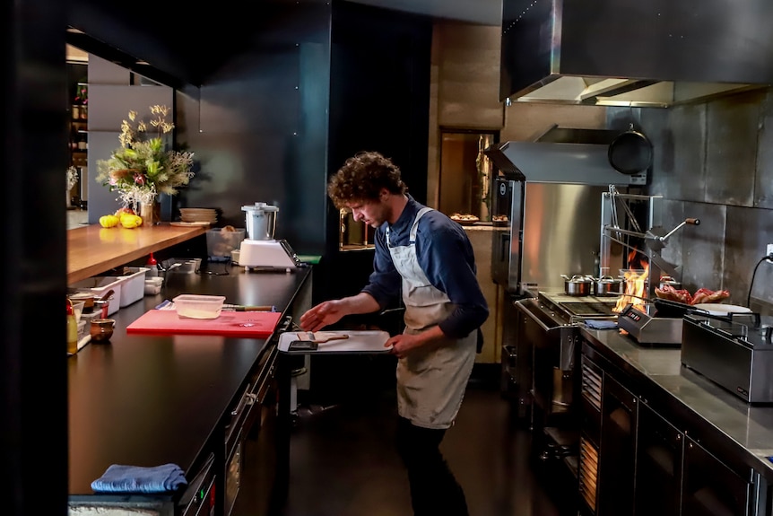 Man wearing apron and blue shirt stands in small professional kitchen with dark coloured stoves and benchtops 