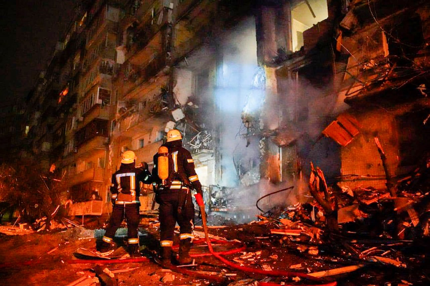 Two firefighters are seen from behind inspecting a badly damaged building lit in a red hue