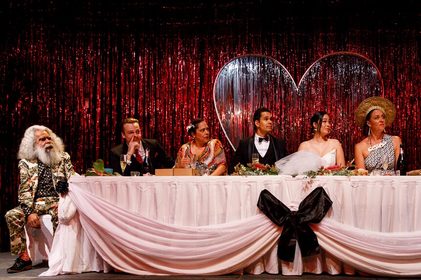 Stage with 8 people seated at wedding reception table and red-metallic mylar curtain behind.