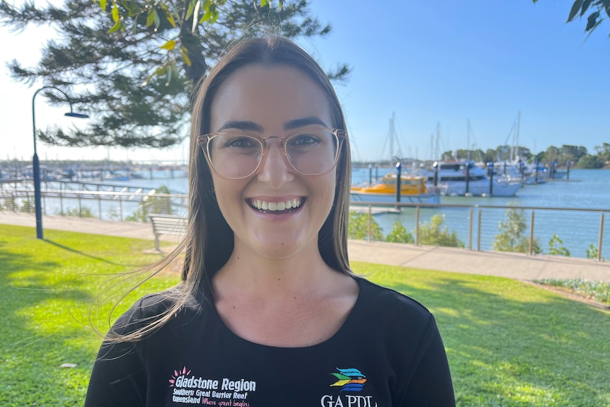 A young woman in black t-shirt, glasses, stands in a park adjacent to a marina smiling at the camera.