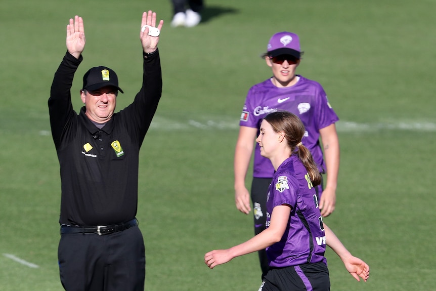 Tayla Vlaeminck walks with her head down past an umpire holding both arms in the air to signal a six.