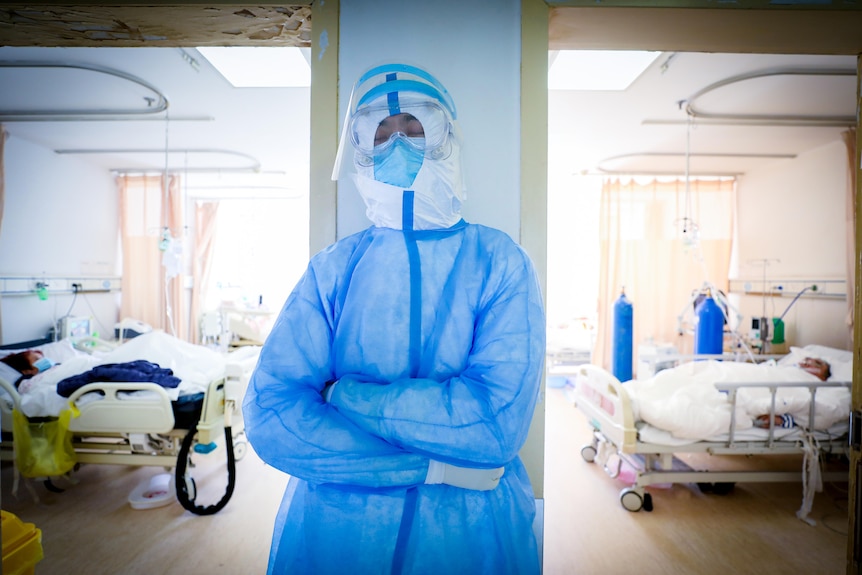 A man in full PPE leans against a pylon between two hospital rooms and closes his eyes 