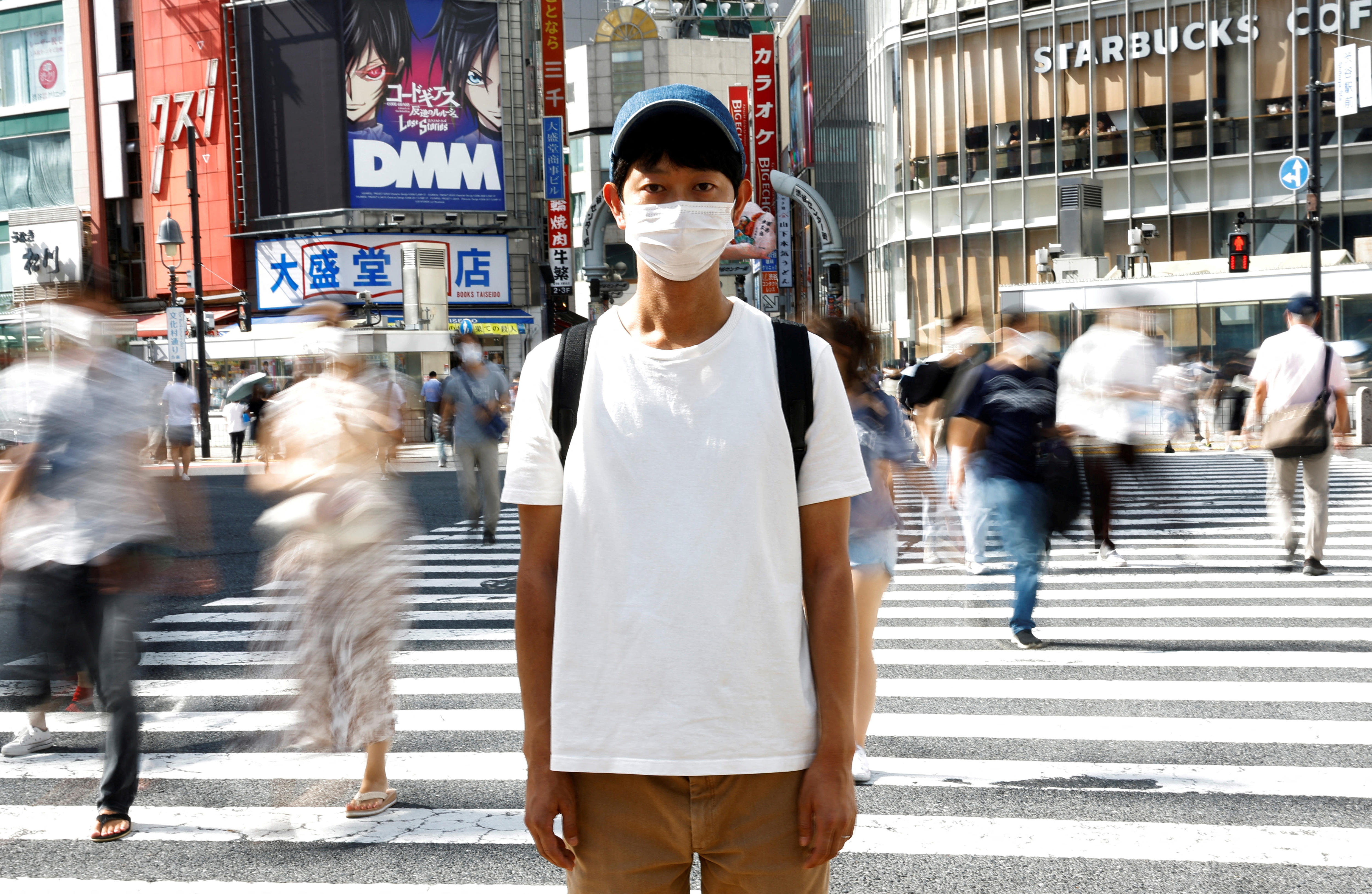 A skinny Japanese man in a white tshirt, baseball cap and mask poses on a busy zebra crossing in Japan. 