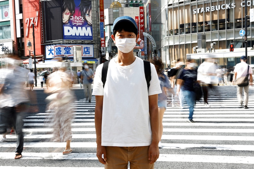 A skinny Japanese man in a white tshirt, baseball cap and mask poses on a busy zebra crossing in Japan. 