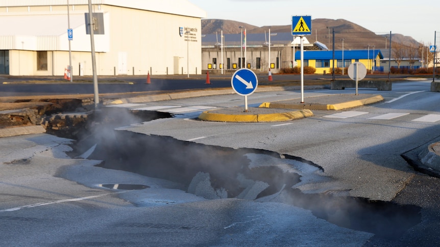 A street is pictured with a very large crack in it from which steam is rising. 