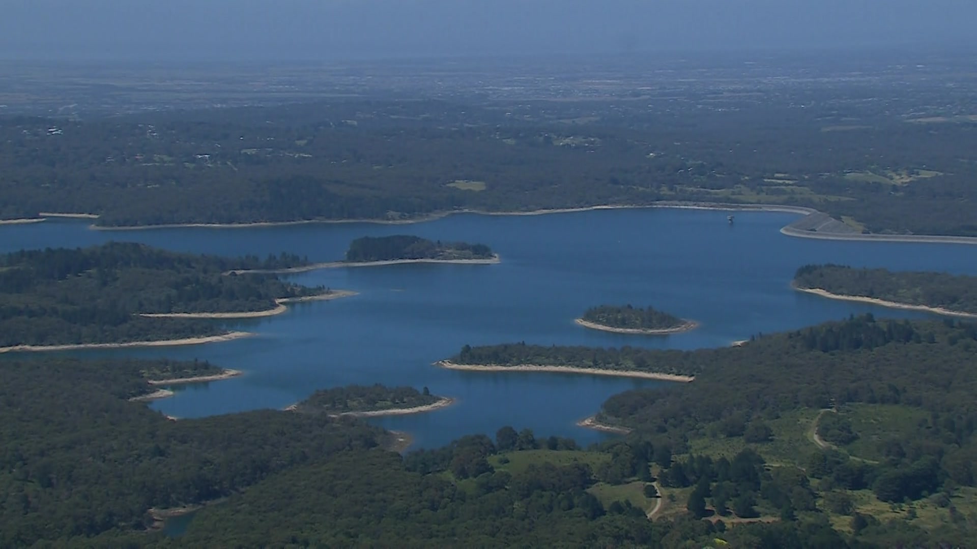 An aerial shot of a large dam surrounded by green trees with islands covered in green trees in the water.