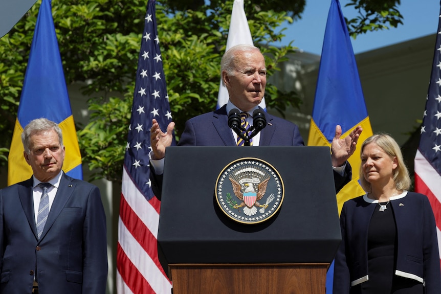 Suited man speaks at lectern with man to his right, and woman to his left.