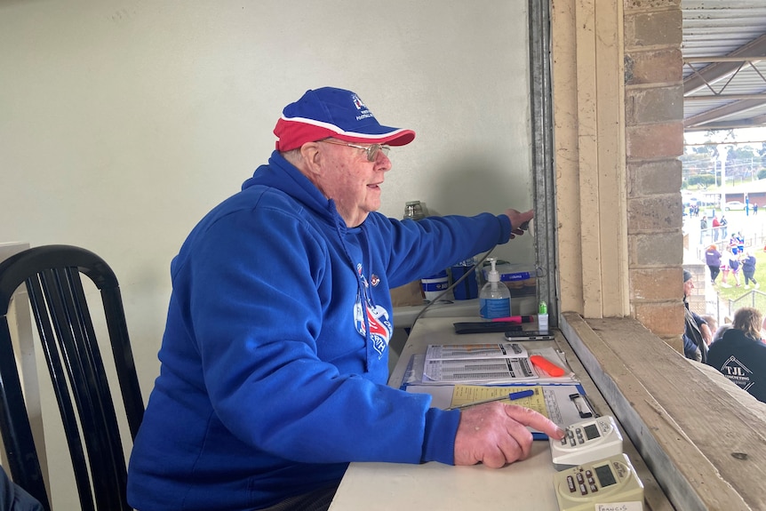 A man in blue and red jumper and hat is looking out a window, with his hand on a stopwatch and the other hand on a siren button