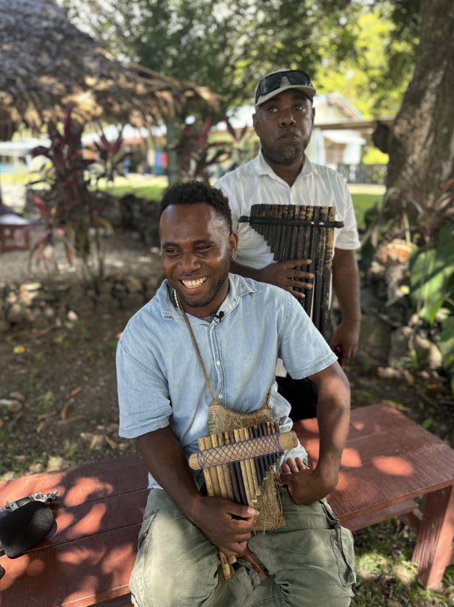 Solomon Island man sits outdoors, wears blue shirt and flute around his neck. Smiles looking to his right