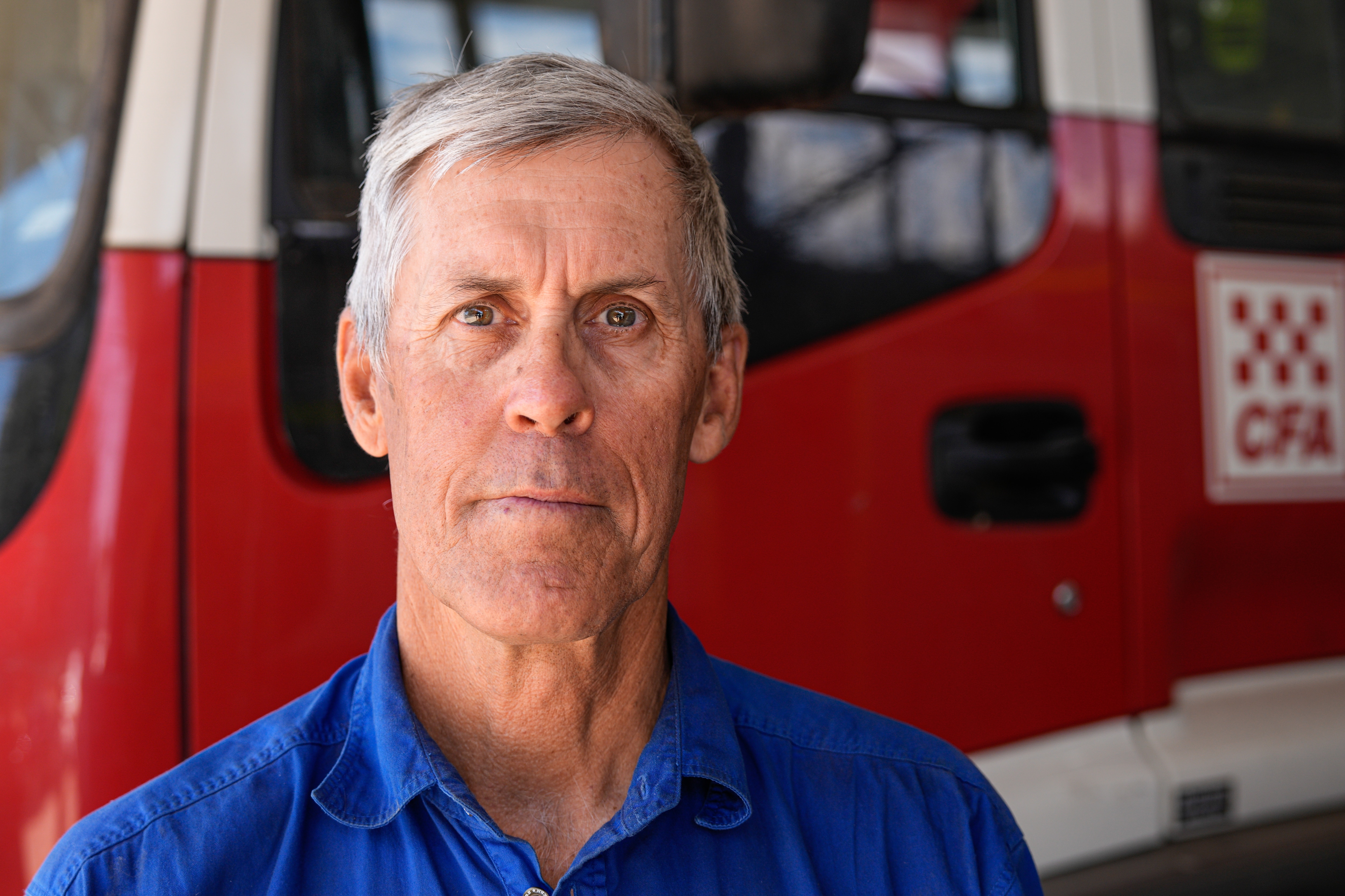 Un hombre con pelo gris y uniforme de bombero se para frente a un camión de bomberos.