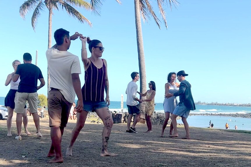 A man and a woman holding hands in a dance pose on a grassy area next to a beach, other dancers around them