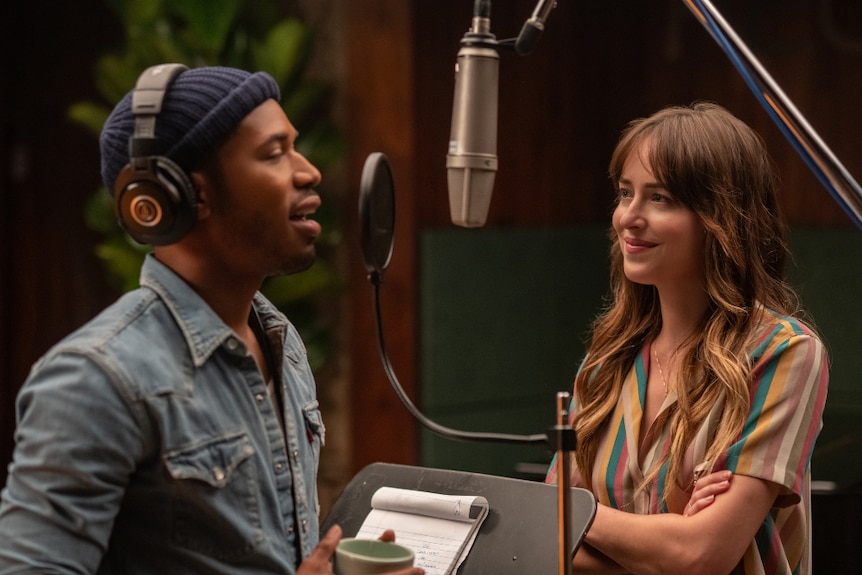 A man wears headphones over navy beanie and stands singing into recording studio mic as a smiling woman watches on close by.