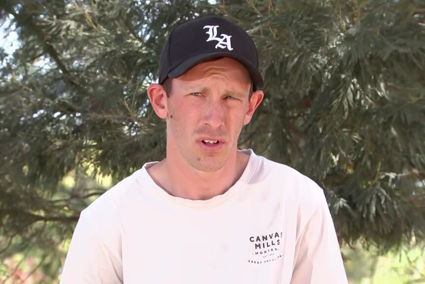 A man wears a white t-shirt and black cap with the letters L A and stands in front of a tree, looking off camera.