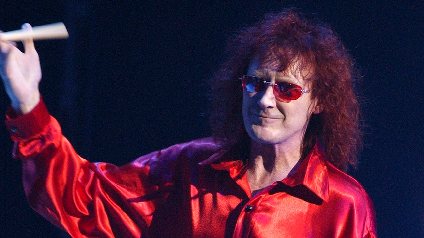 A portrait orientated photo of a man with curly hair and glasses spinning some drum sticks during a concert