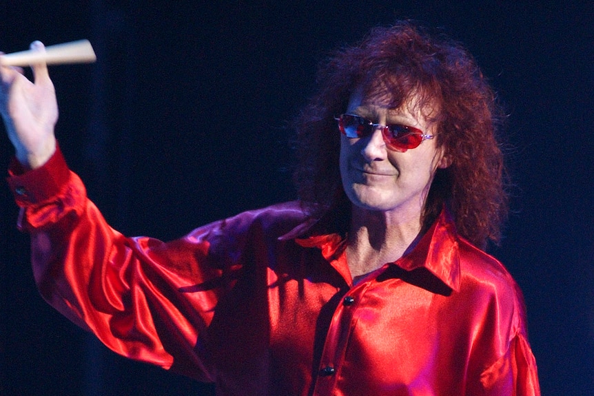 A portrait orientated photo of a man with curly hair and glasses spinning some drum sticks during a concert
