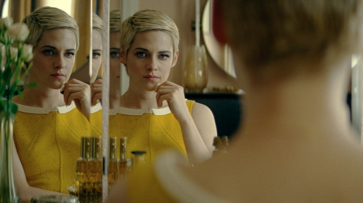 A woman with short blonde hair in yellow 1960s dress and hand touching chin looks at multiple reflections of herself in mirror.