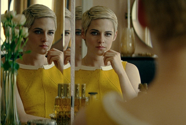 A woman with short blonde hair in yellow 1960s dress and hand touching chin looks at multiple reflections of herself in mirror.