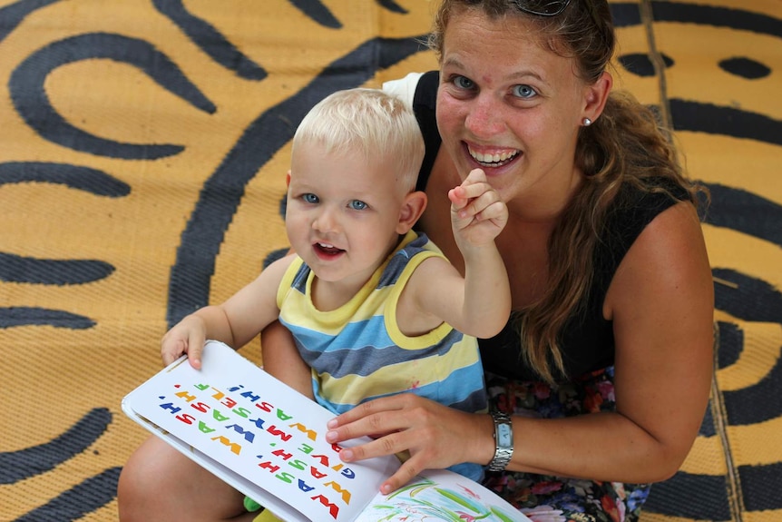 A toddler boy sits on the lap of a woman as they read a book