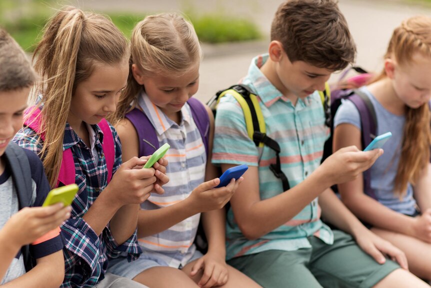 Five primary school students sit side-by-side on a wall and use mobile phones.