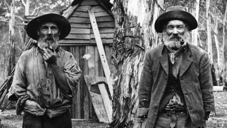 Archive photo of two cedar-getters smoking pipes and standing in the forest in front of a wooden shack.