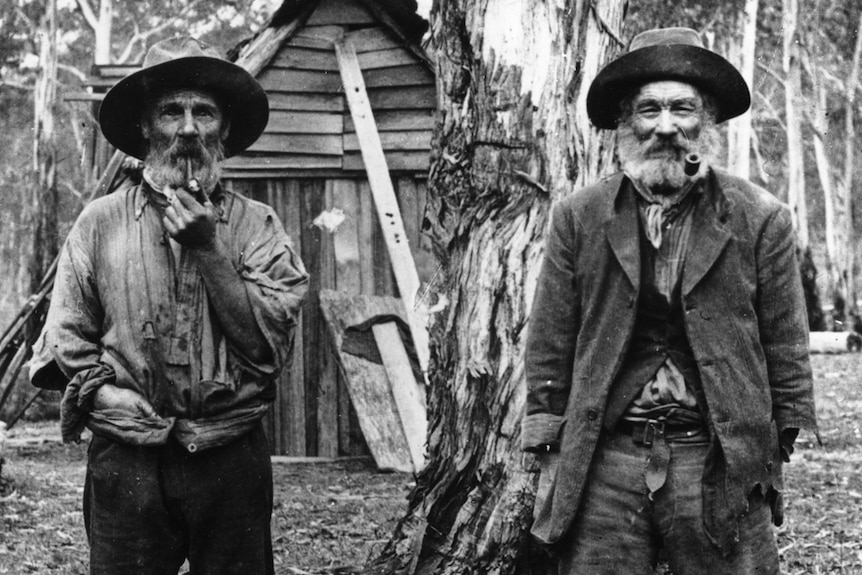 Archive photo of two cedar-getters smoking pipes and standing in the forest in front of a wooden shack.