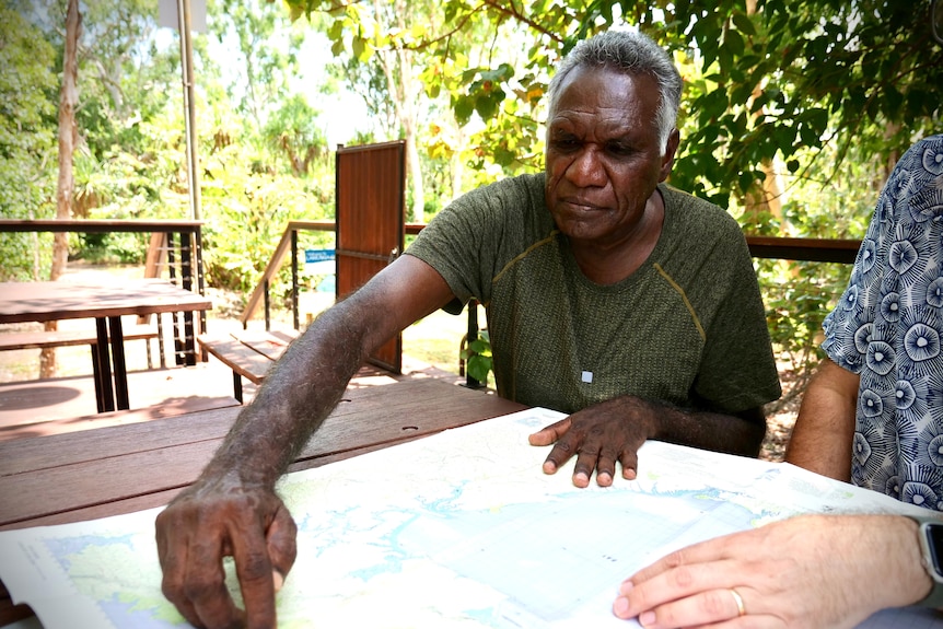 A man with dark skin and white hair sits outside pointing at a map spread on table.