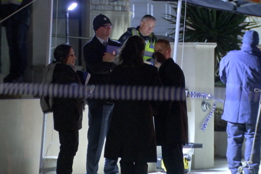 A uniformed officer and a number of police in plain clothes stand outside a house behind police tape.