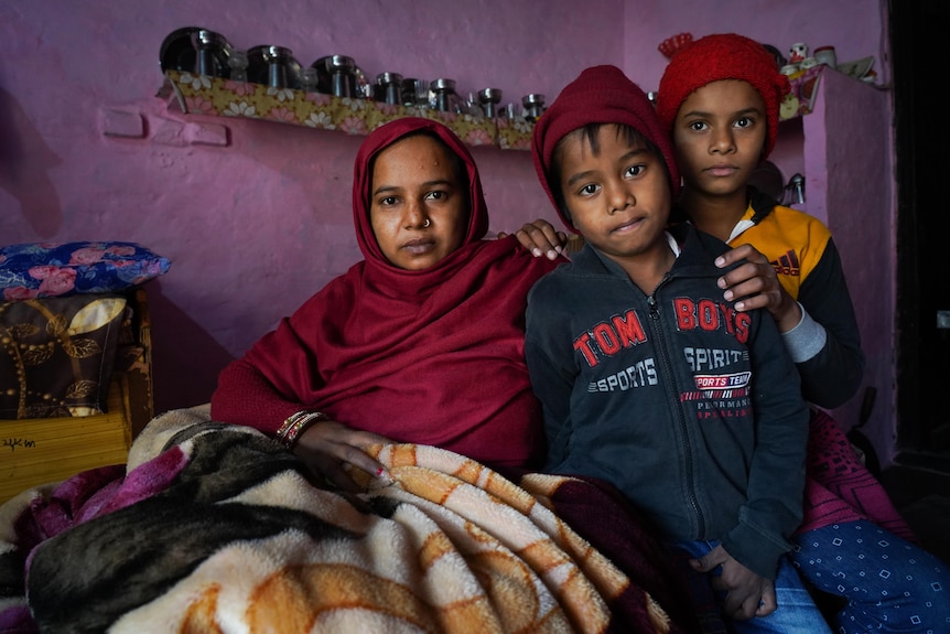A woman wearing a maroon headscarf sits on a bed with her two children, inside a room with pink walls