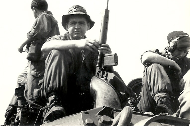 Laurie sitting on top of a tank holding a gun.